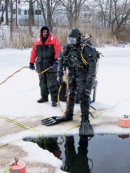 A tech diver stands on the edge of an icy pool next to a man in a red coat