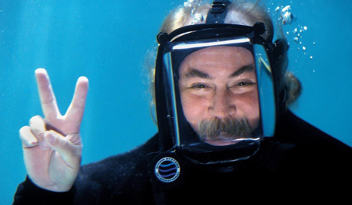 Man in scuba mask, and has a mustache, is giving the peace sign