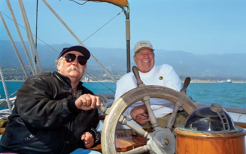 Two men sit on a boat behind ship's wheel. Both men are wearing hats, and the mustached man is wearing black aviator sunglasses.