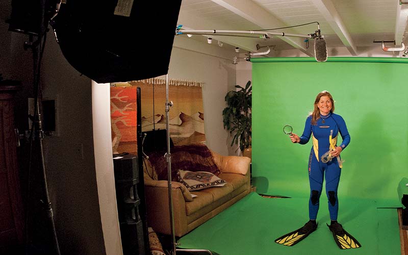 Blonde woman in blue wetsuit and flippers, stands in front of green screen