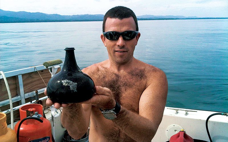 A shirtless man wearing sunglasses, is standing on a boat and is holding a black, old bottle