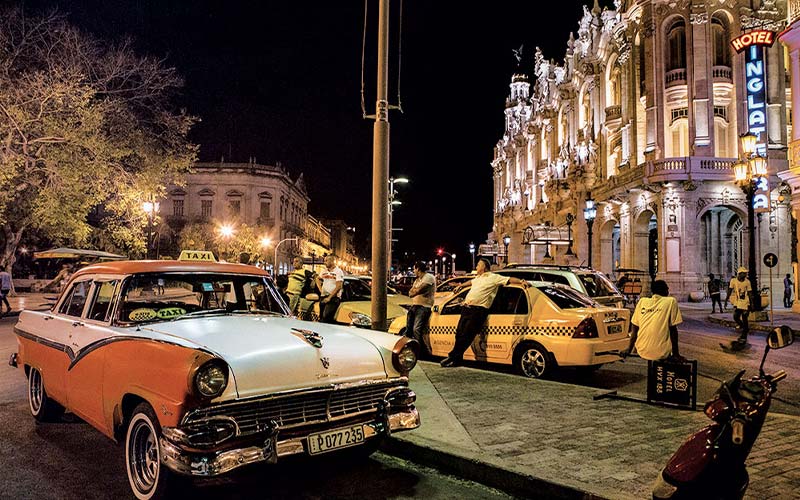 A charming street in Havana at night. There are taxi cabs and an antique car on the road