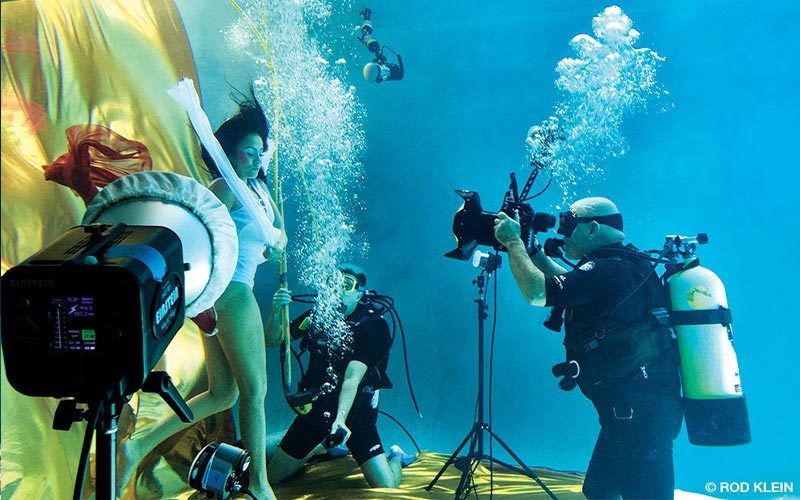 A scuba diving photographer is behind the camera shooting a model who is holding her breath