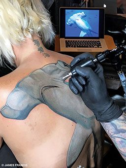 A blonde woman gets a hammerhead tattoo on her back