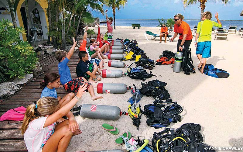 A group of children sit on a walkway, with their hands raise, taking a scuba diving class