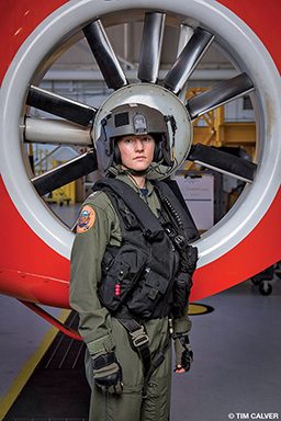 A person wears a US Coast Guard uniform and stands in front of a jet engine