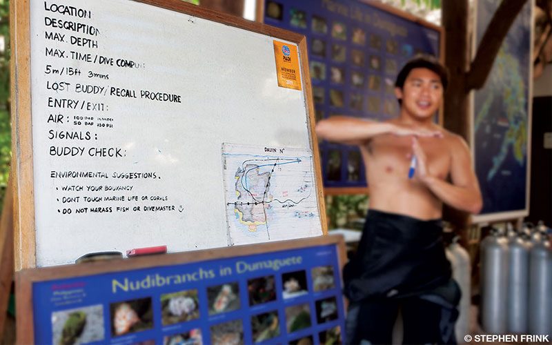 A shirtless man stands in front of a white board and instructs a group of an upcoming dive