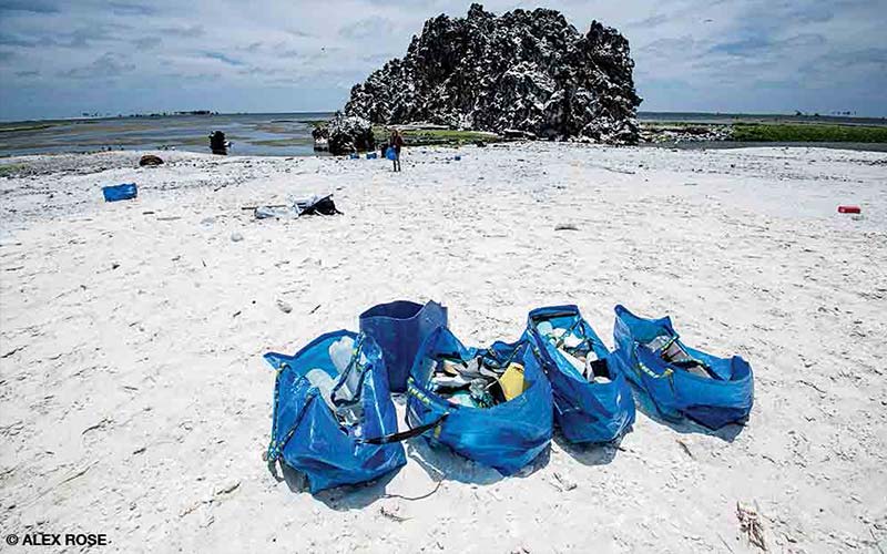 Several large blue bags rest on a clean, sandy beach. The bags are full of trash picked up off the beach