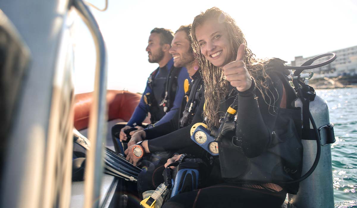 Female diver sits on the edge of a boat and gives a thumbs up to the camera. In the background are two male divers.