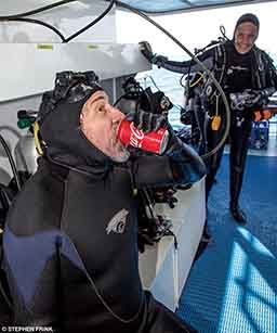 A man chugs a can of Coke and a woman laughs at him in the background. Both are in scuba gear.