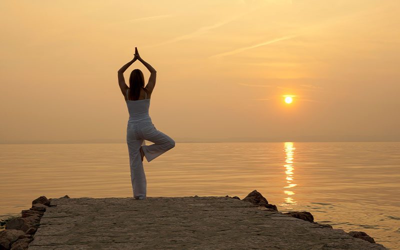 A woman stands at the water's edge performing yoga. It is sunset