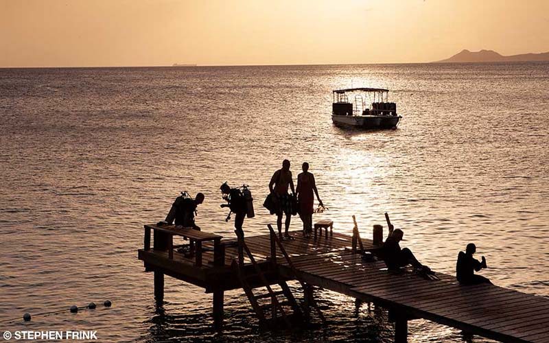 A group of divers dawdle on a dock at sunset. A boat is in the background.