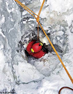 A person in a red helmet searches for a new cave entrance. They are surrounded by ice and snow.