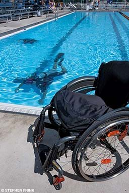An empty wheelchair is on the pool deck while its owner dives in the pool nearby. 