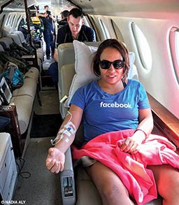 A woman in sunglasses is on a plane. A medical crew is behind her and she has an IV in her right arm.