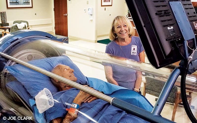 An old man is in a hyperbaric chamber. He's accompanied by a woman in a purple shirt.