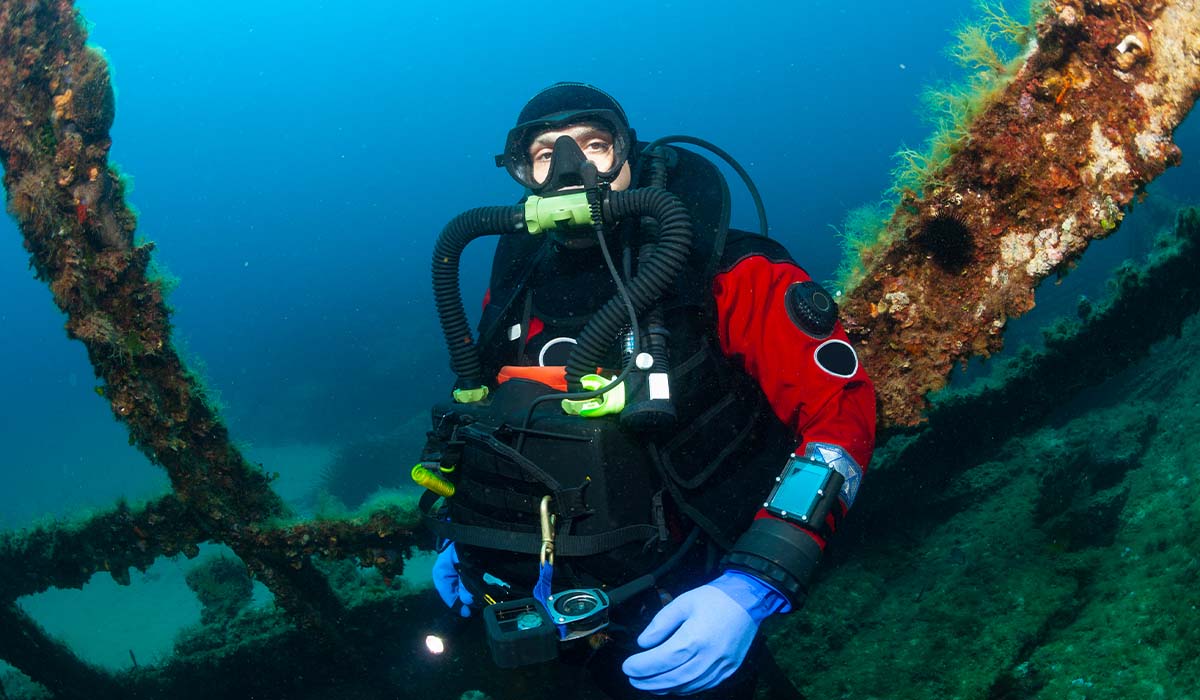 A rebreather diver, in a red suit, floats near a wreck