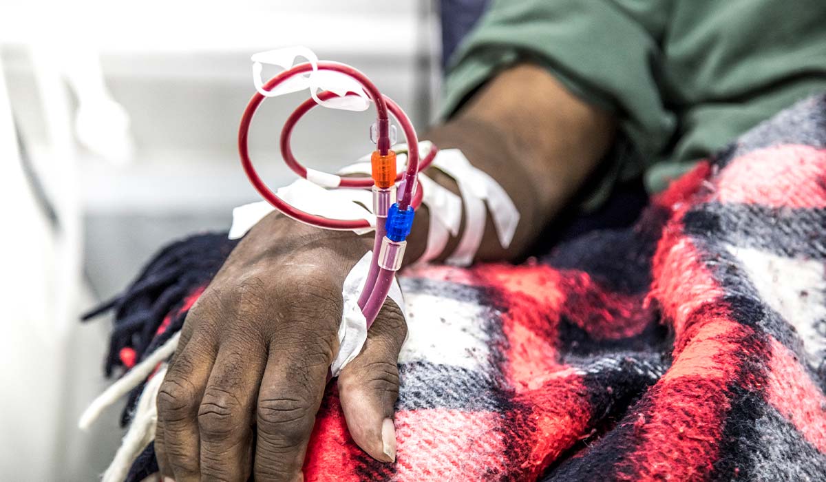 The right hand of a Black man is hooked up to tubes and is receiving dialysis. His lap is covered with a red plaid blanket