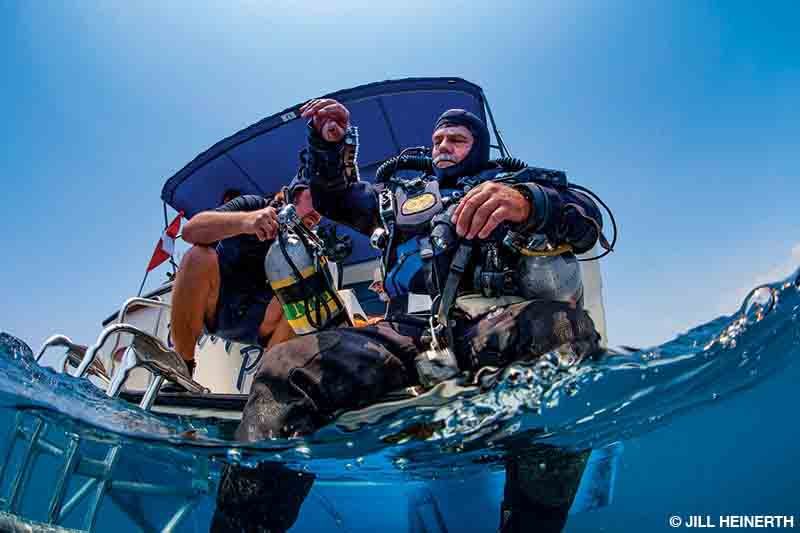 A man on a boat helps a diver with a scuba cylinder before the diver enters the water.
