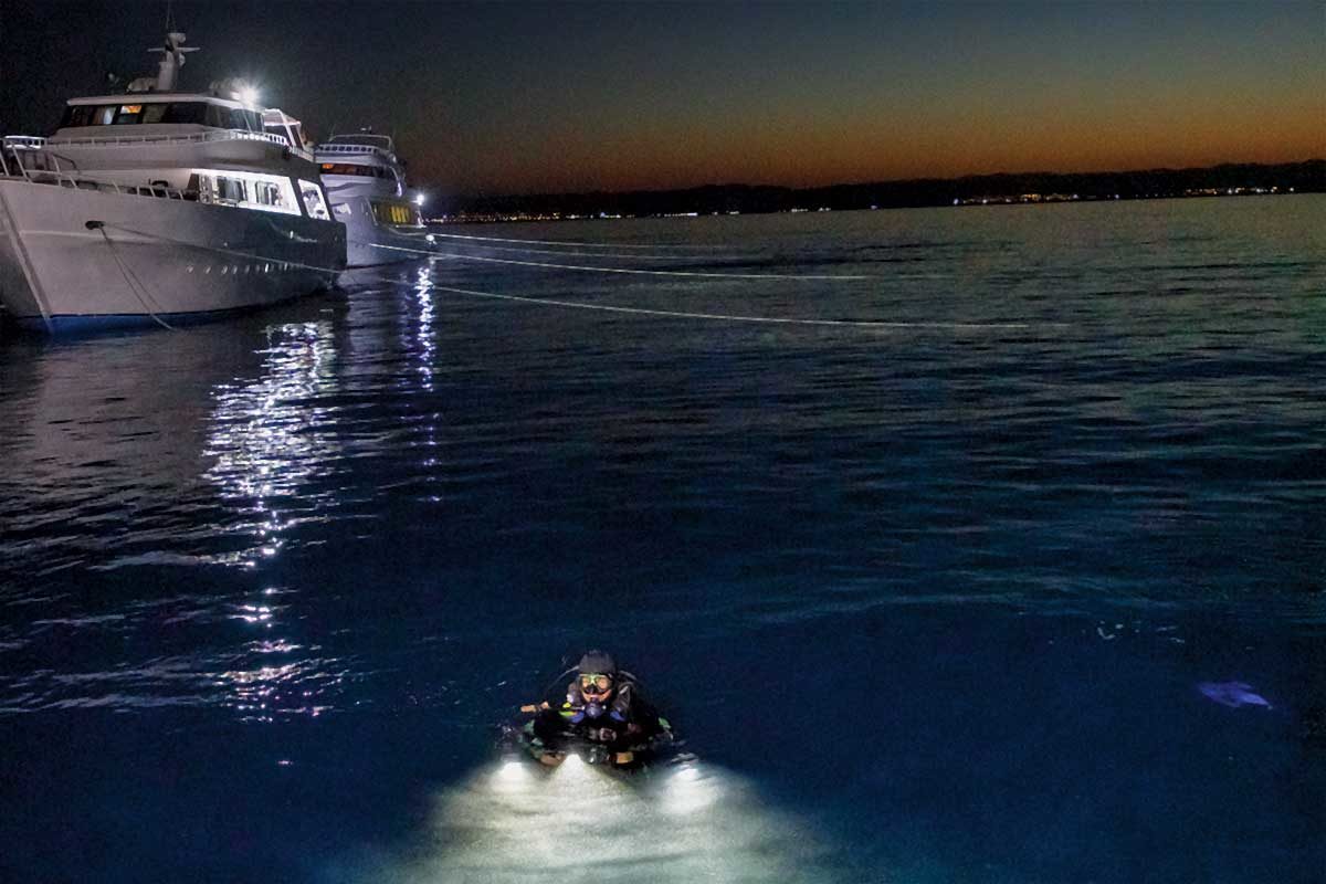 A diver is in the water at the surface at dusk as a liveaboard waits nearby.