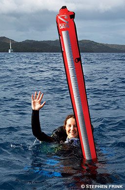 A female diver with a deployed surface marker buoy waves at the surface.