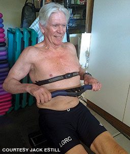 A shirtless older man exercises on an indoor rower while wearing a heart monitor.