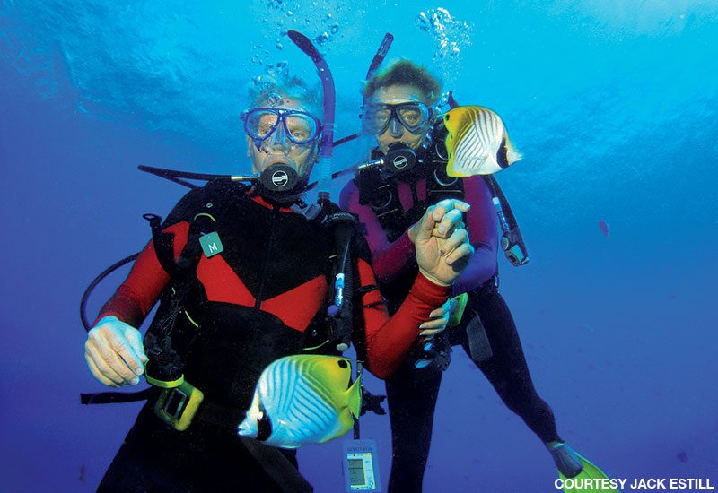 Male and female diver in red and black wetsuits pose for a photo underwater before descending