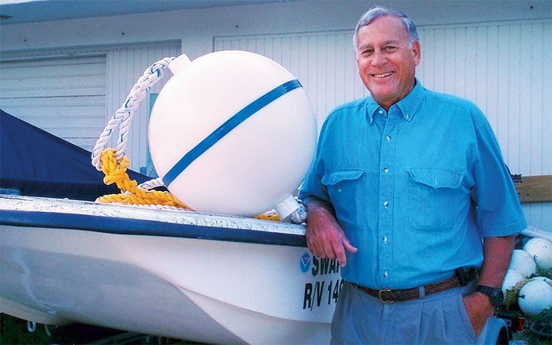 A man in a blue shirt, John Halas, stands next to a boat with a mooring buoy on top.