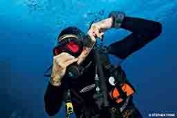 A diver tries to equalize his ears by clutching his nose. He's wearing a dive hood.