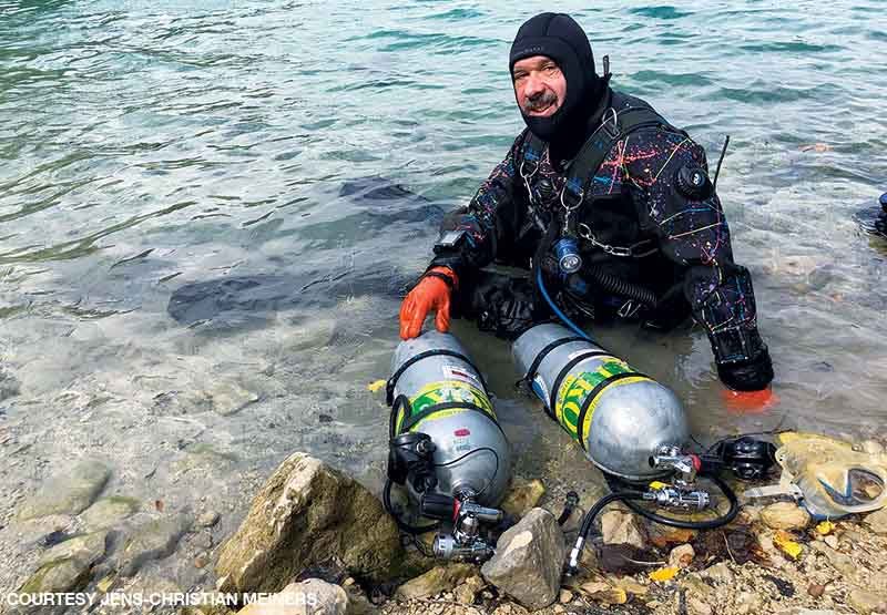 Meiners in a drysuit sits in the water near shore after a dive with two nitrox tanks