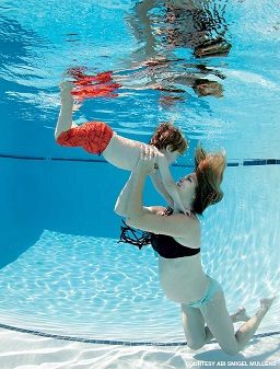 A pregnant woman in a bikini holds a child wearing red swim trunks. They are both submerged in a swimming pool.