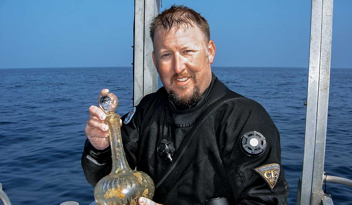 Bearded man in scuba suit stands on boat and holds an artifact
