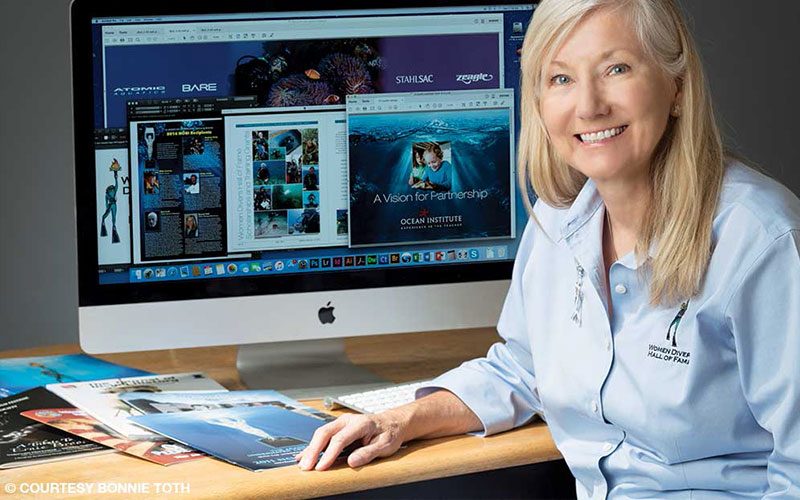 A blonde woman sits in front of a Mac computer and smiles to the camera