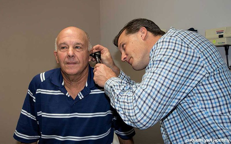 A doctor in a blue plaid shirt, examines the left ear of a bald man