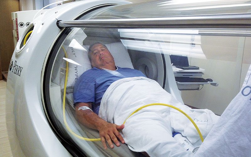 A sad-looking man rests in a hyperbaric chamber. He's torso and legs are covered by a white blanket and he's wearing a blue hospital gown.