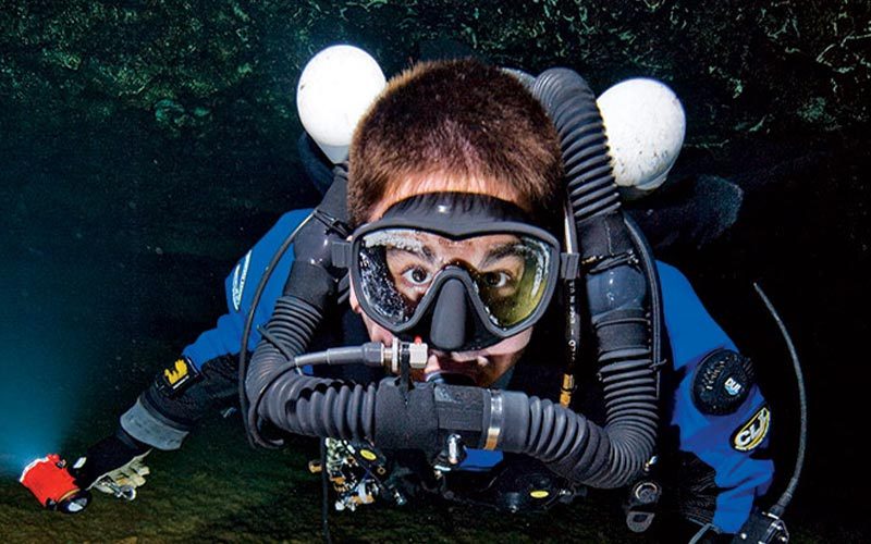 A rebreather diver poses in a cave and wears a blue and black wetsuit