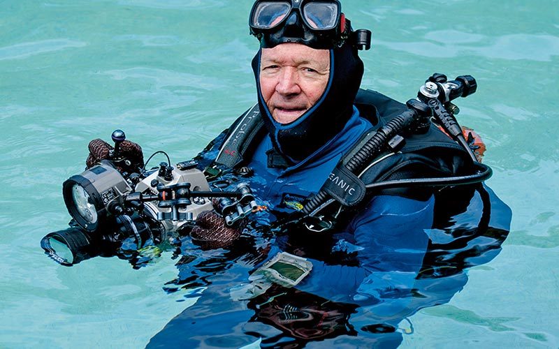 Roger Steene wears a drysuit and holds a camera, while bobbing in the water
