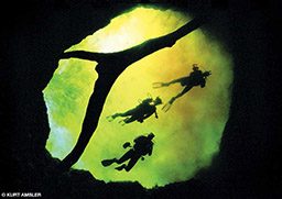 Three cave divers swim through an eerie-looking cave. The light appears lime green