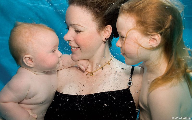 Zena Holloway (center) holds a baby in her right arm and a toddler in her left arm. All three are submerged underwater.
