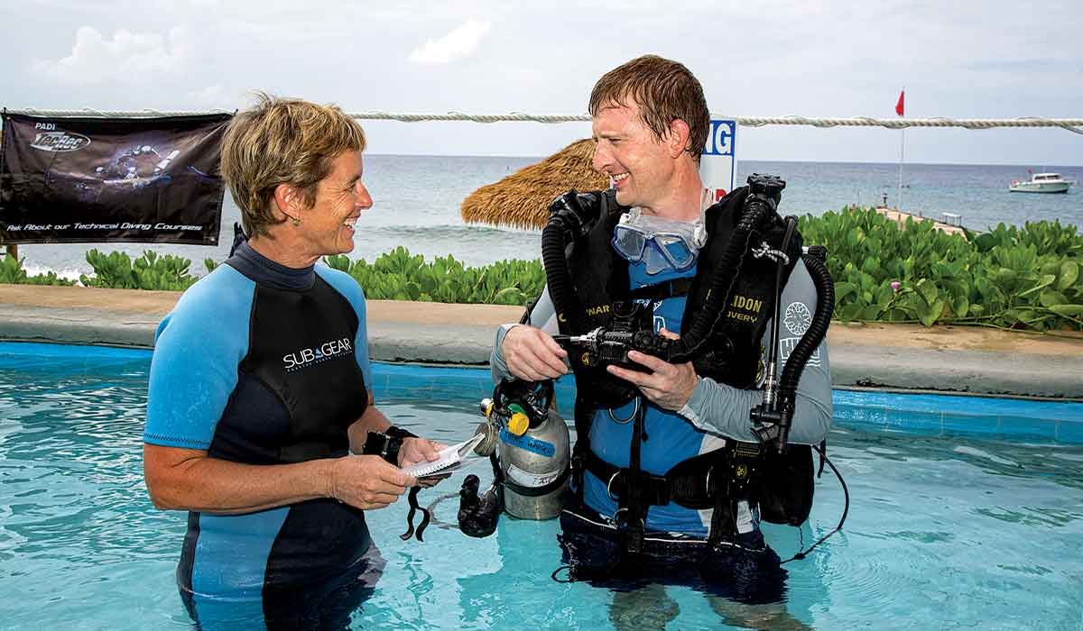 A man in rebreather gear stands next to a woman, in waist-deep water