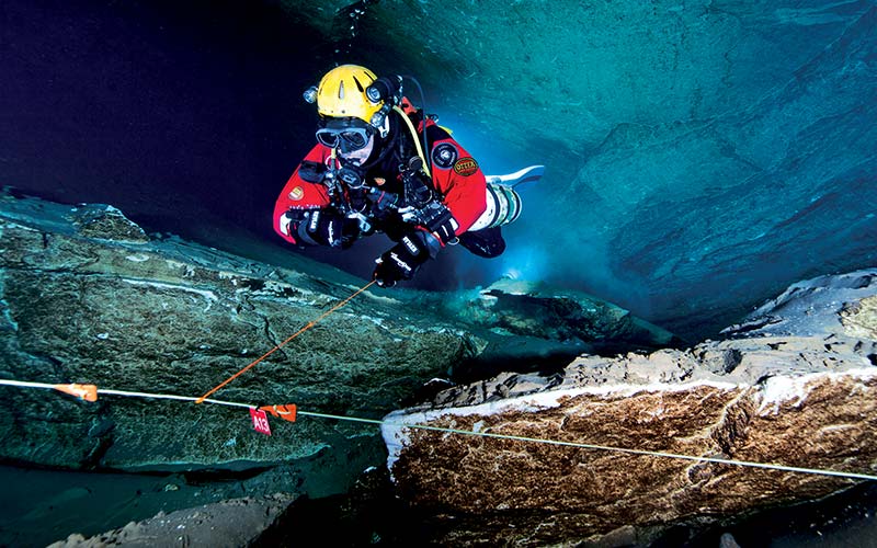 Cave diver in red drysuit and yellow helmet swims through a cave