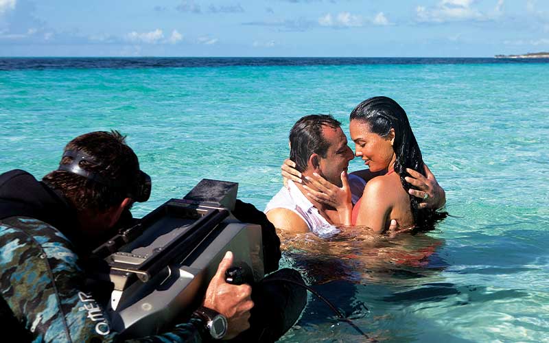 Couple embraces in the shallows of tropical waters, a cinematographer is nearby filming them