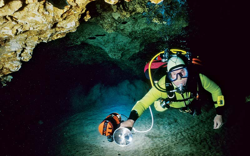 Diver in neon-green wetsuit floats through a cave holding a lit flashlight