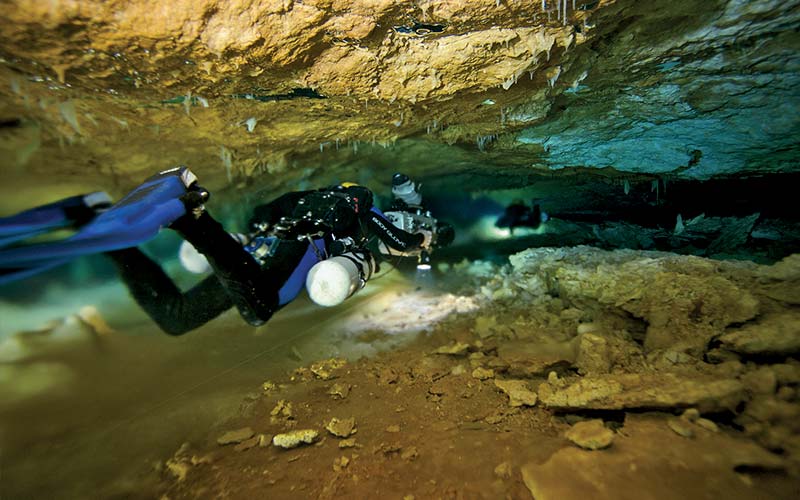 Diver swims through a thin slit in a cave holding a light