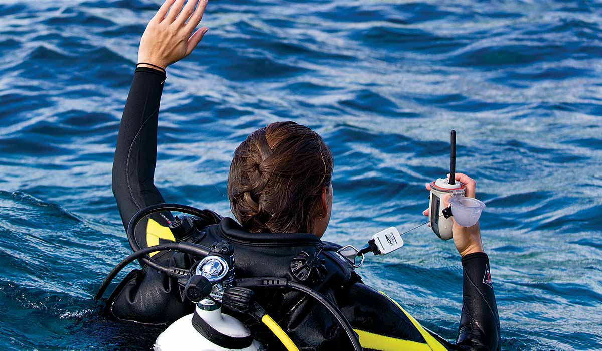 Female diver holds signaling device and is also waving down a boat