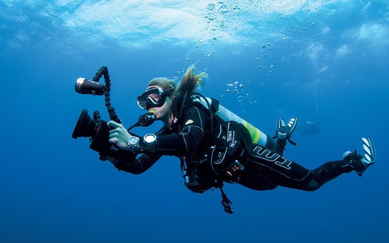 Female diver swims through water holding a huge camera