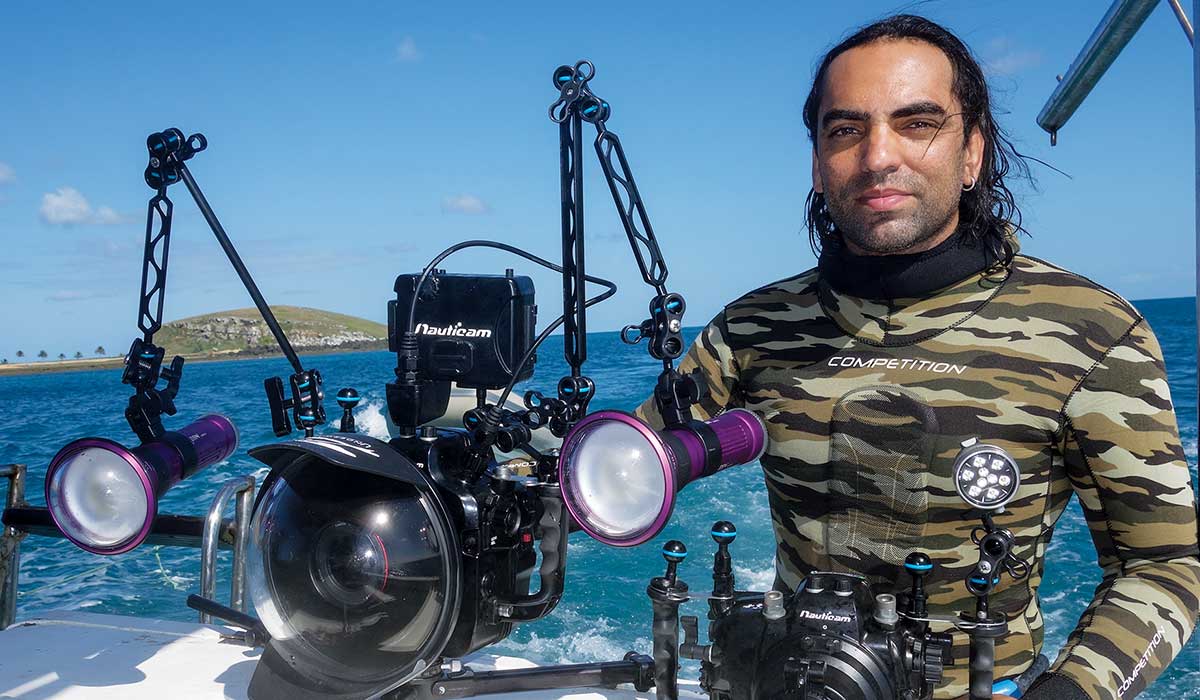 Filmmaker poses on a boat, wearing a wetsuit, holding a camera