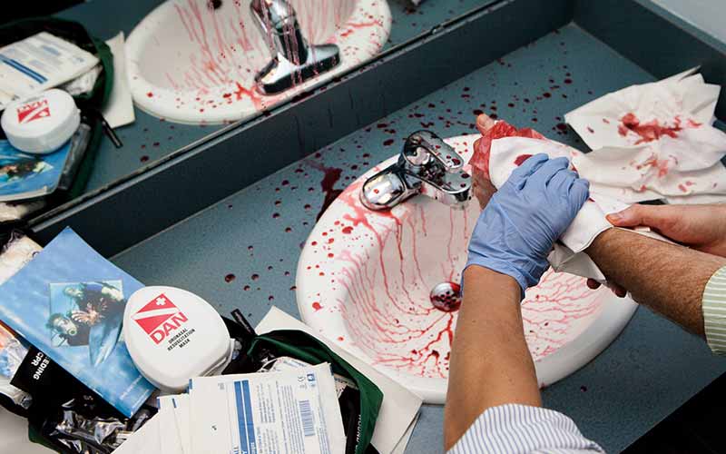 Gloved paramedic hands aid a bloodied hand over a blood-covered sink