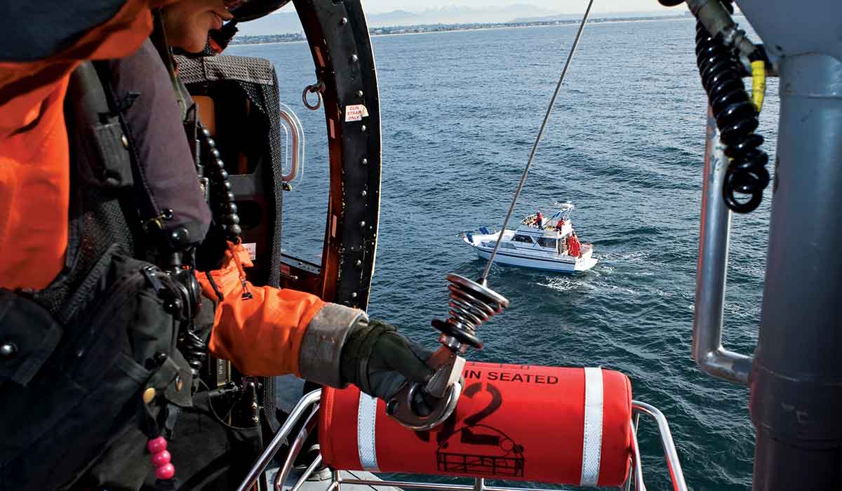 Helicopter crew member works with a hoist. The helicopter is above the ocean. A boat is in the background