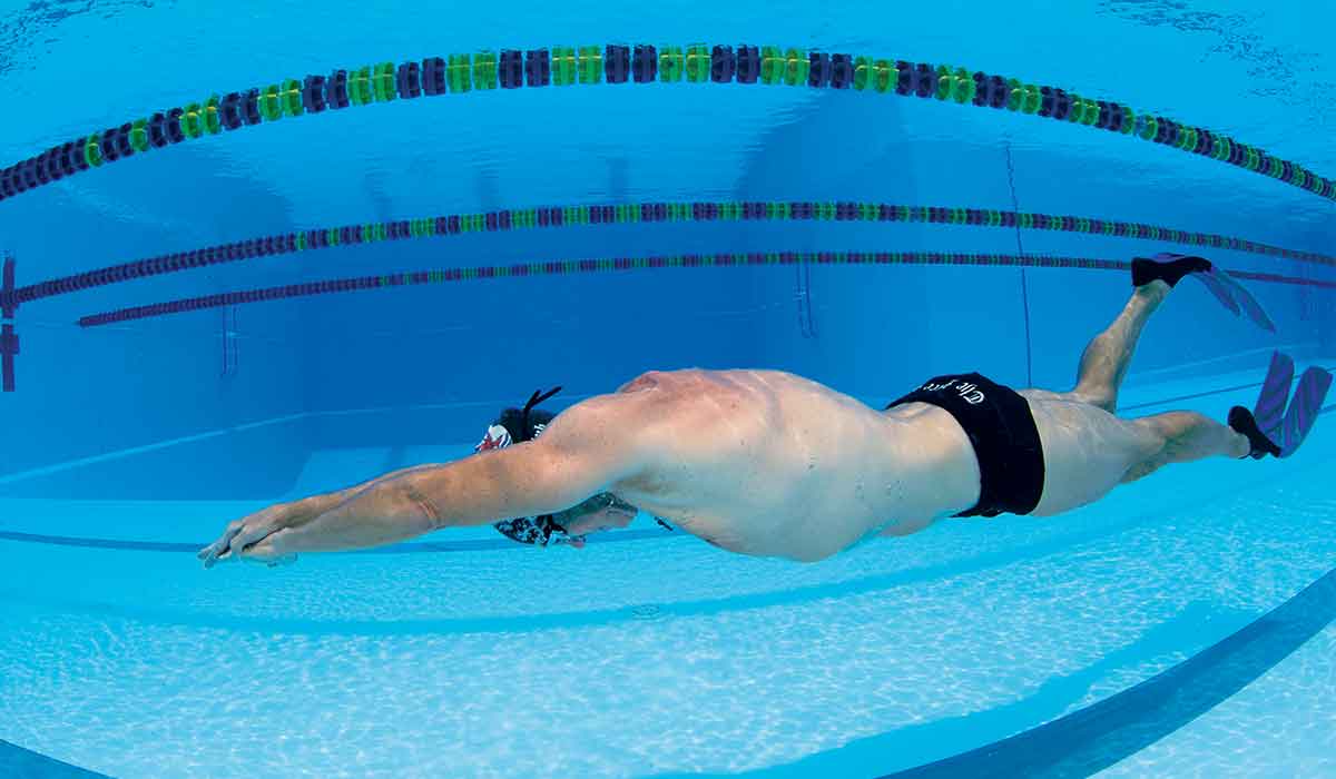 Man in Speedo and blue fins swims underwater in a pool